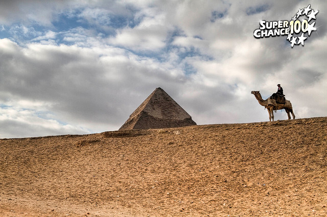 Photographie d'une paysage désertique avec un homme à dos de chameau devant une pyramide.